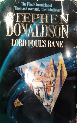 The cover of Lord Fouls' Bane by Stephen Donaldson, The First Chronicles of Thomas Covenant, the Unbeliever. The cover is set in space, a glass or window like pyramid depicts a scene inside of buildings carved out of mountenous rock, with waterfalls and greenery in the foreground. The floating pyramid floats in space with arcs of light and stars flowing behind it.